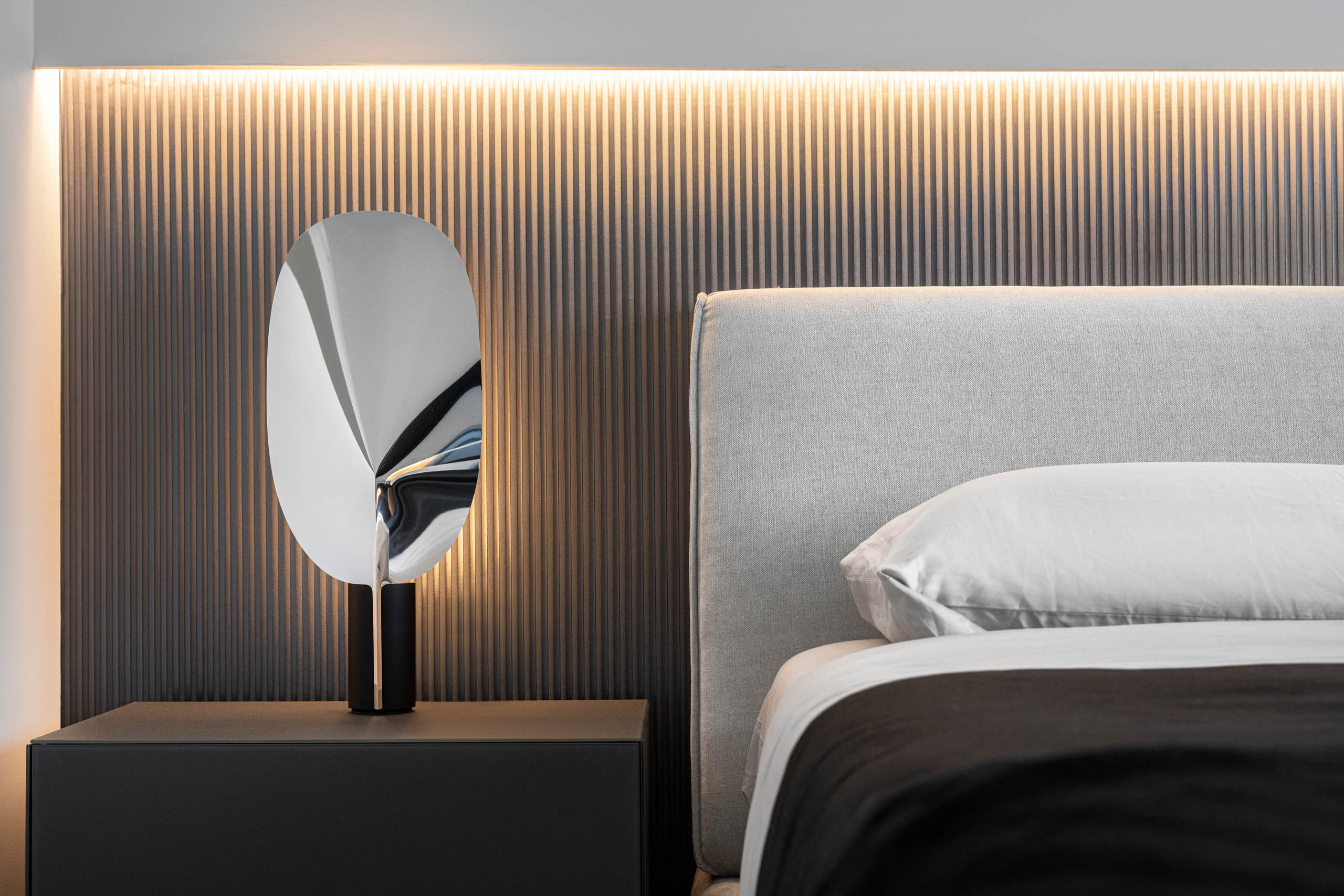 Example of Miami Interior Design project all black and white with white headboard bed, black duvet cover, biomimetic table lamp in leaf format in glossy chrome material. background of stripped black wallpaper with LED cove ilumination.
