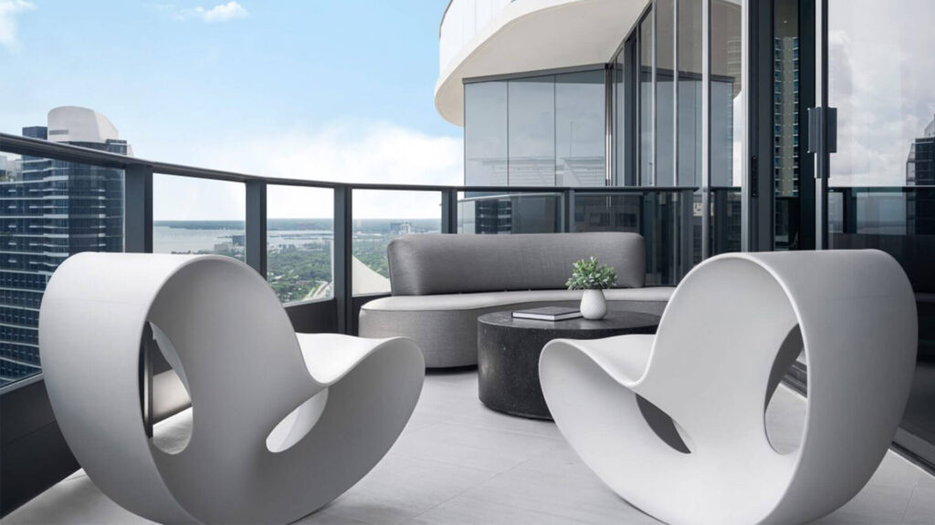 Balcony furniture circular arrangement with organic shape gray sofa, round black terrazzo coffee table and 2 Organic shaped and futuristic white chairs. Background view of Miami and blue sky.