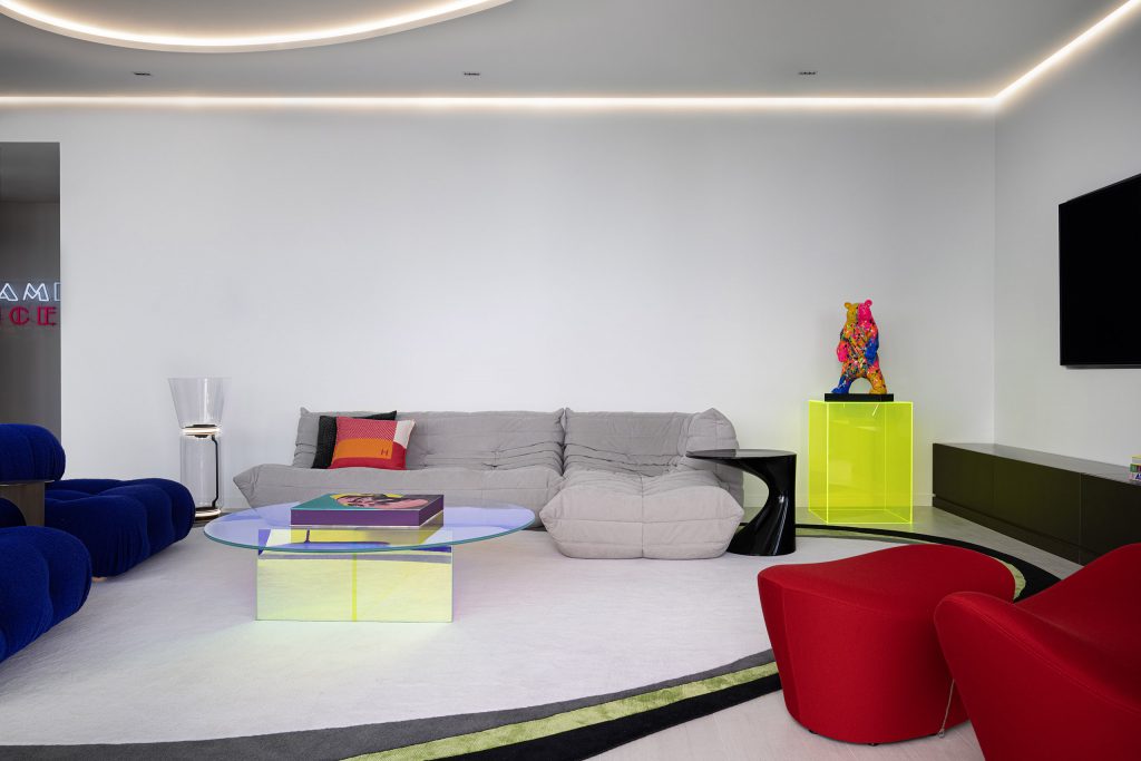 Modern interior design with gray sofa, blue lounge chair, neon yellow pedestal with sculpture, and red accent chairs.