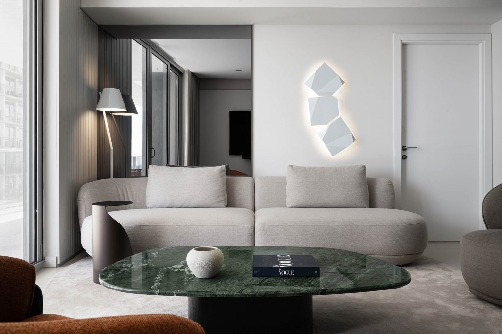 Curved sofa and green stone coffee table in the living room of FM Apartment with illuminated wall art and floor lamp.