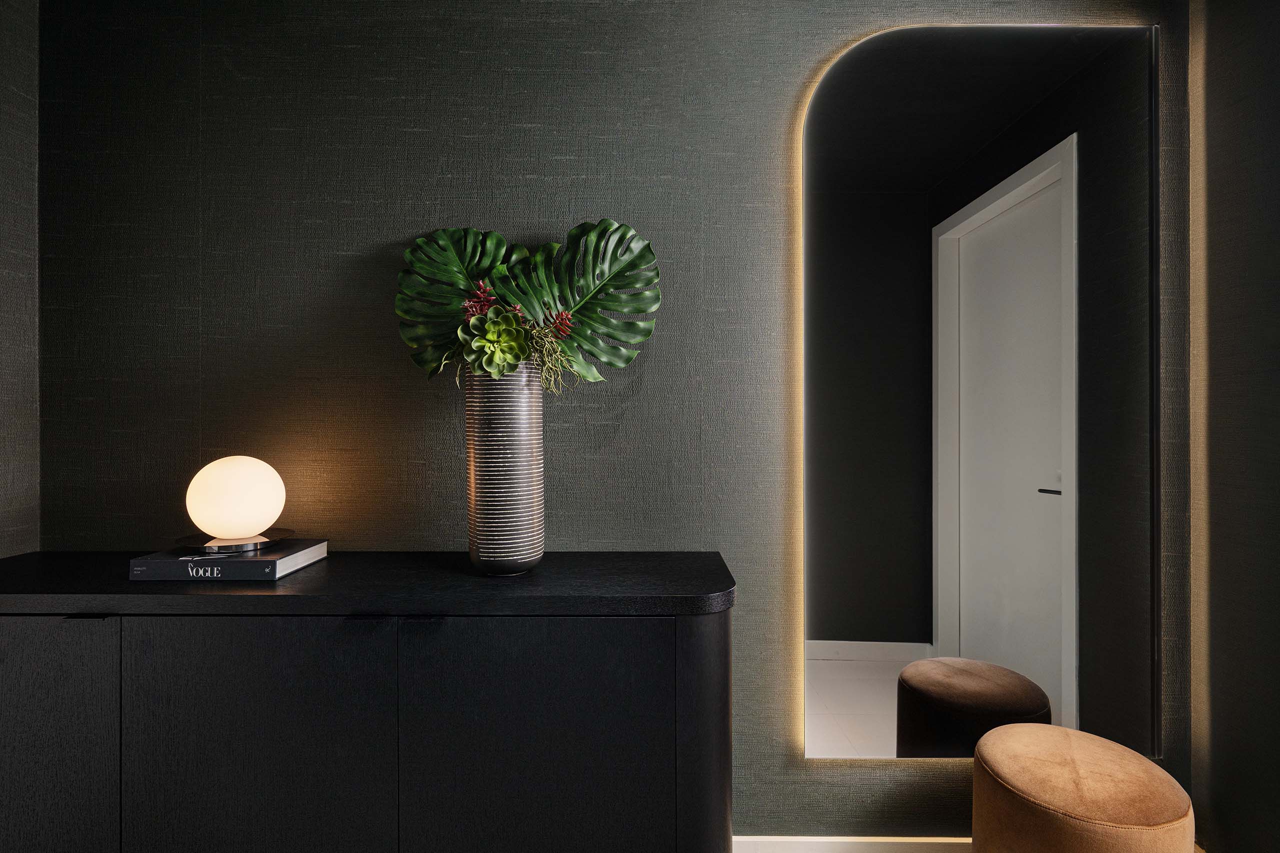 Entry console in FM Apartment with illuminated arched mirror, dark cabinetry, and floral arrangement.