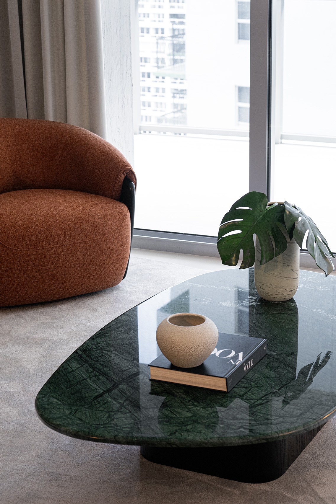 Green stone coffee table detail in FM Apartment with ceramic vessel, plant, and rust lounge chair by the window.