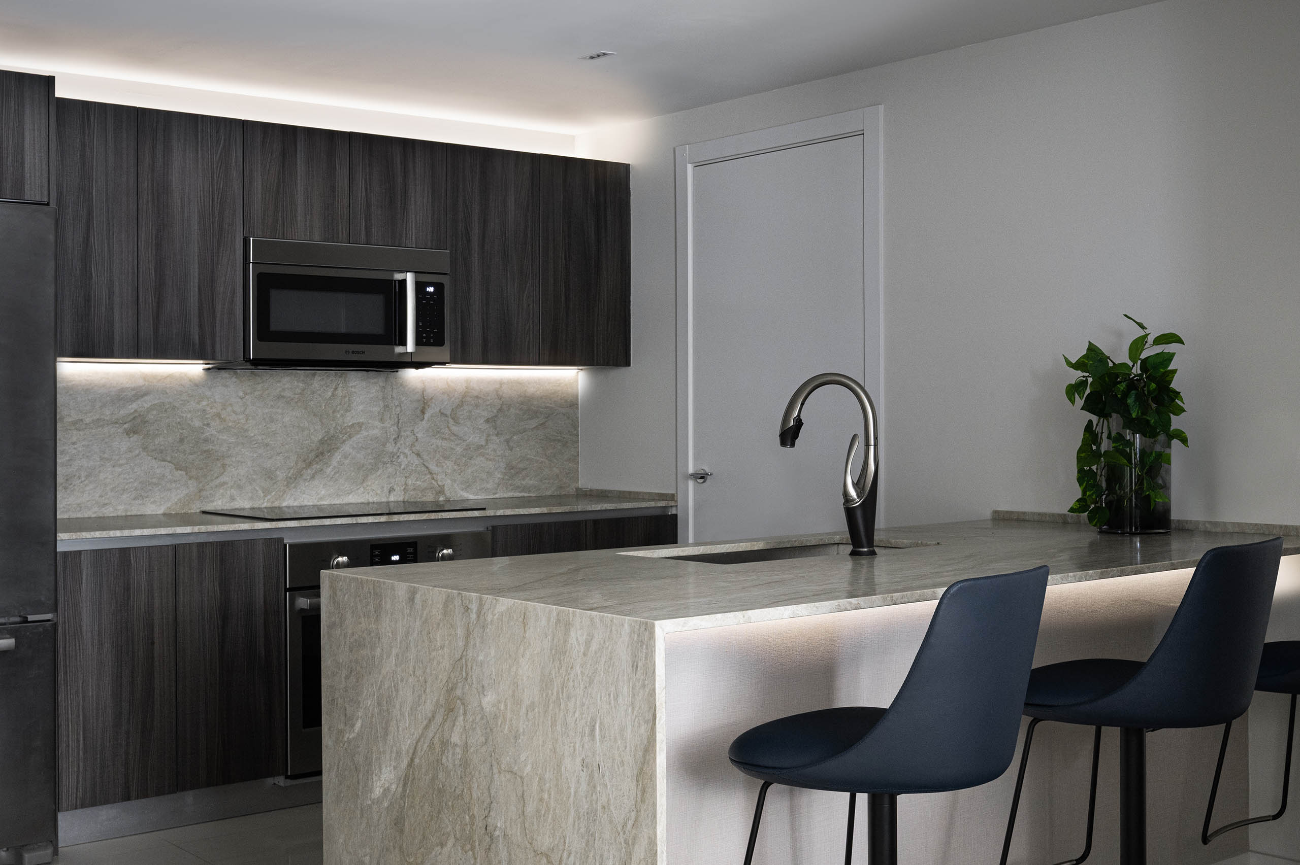 Kitchen in FM Apartment with light stone waterfall island, dark wood cabinetry, and integrated lighting.