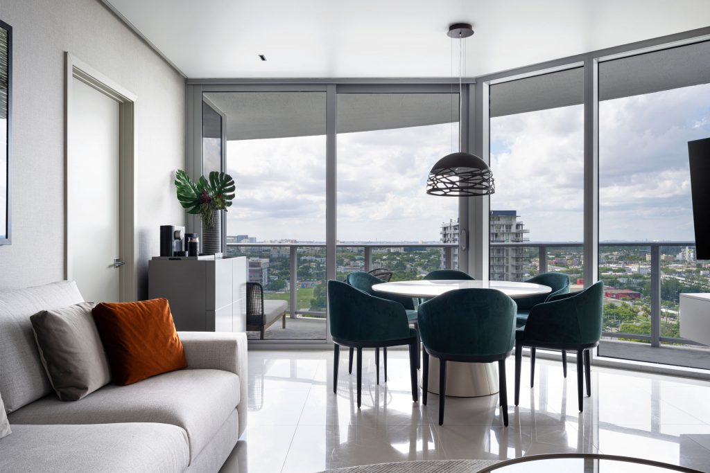 Dining corner in MB Apartment with round white table, green velvet chairs, pendant light, and skyline views through corner glazing.