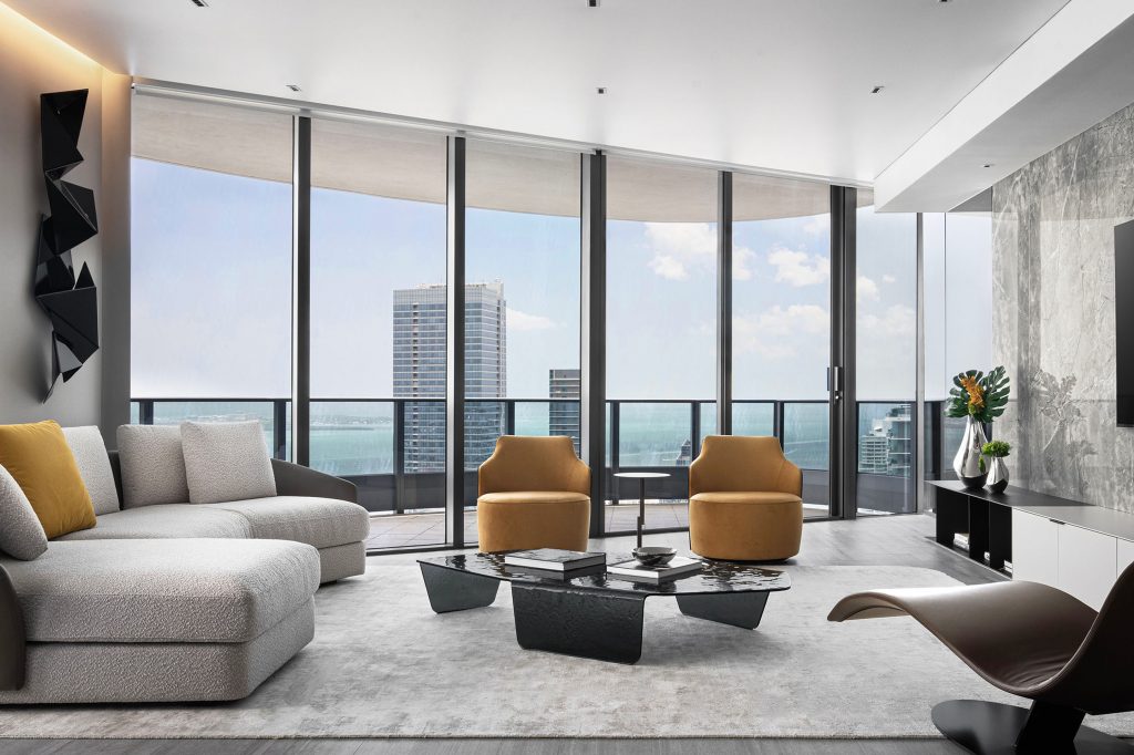 Living room at NR Residence in Brickell Flatiron with bouclé sectional, mustard swivel chairs, smoked-glass coffee table, and Biscayne Bay views.