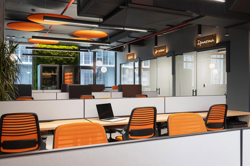 Open office seen from behind orange-backed task chairs, with rows of desks facing glass meeting rooms labeled “Zaxy,” “Rider,” and “Ipanema,” and acoustic panels above