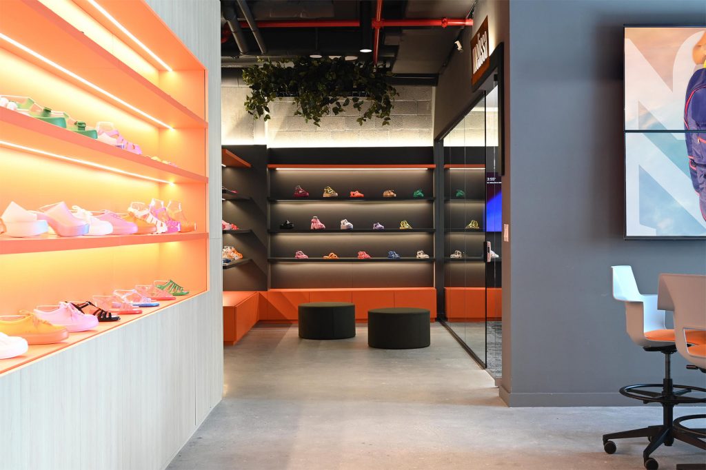 Office interior with brightly lit orange shelving displaying rows of shoes on the left, a corridor leading to a darker display wall with additional footwear, and a glass partition revealing a meeting area with chairs on the right