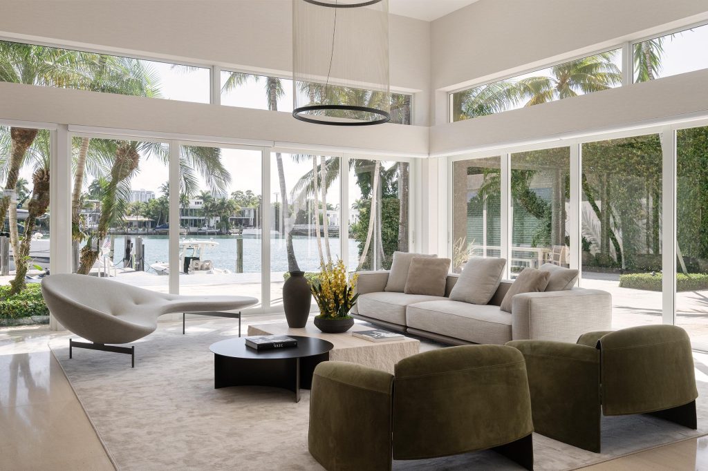 Living room corner with clerestory windows and full height glazing onto palms, boats, and water, a curved white chaise on metal legs, a pale sofa with taupe cushions, two olive suede armchairs, and a stone coffee table with a dark vase and yellow orchids beside a round black metal table.