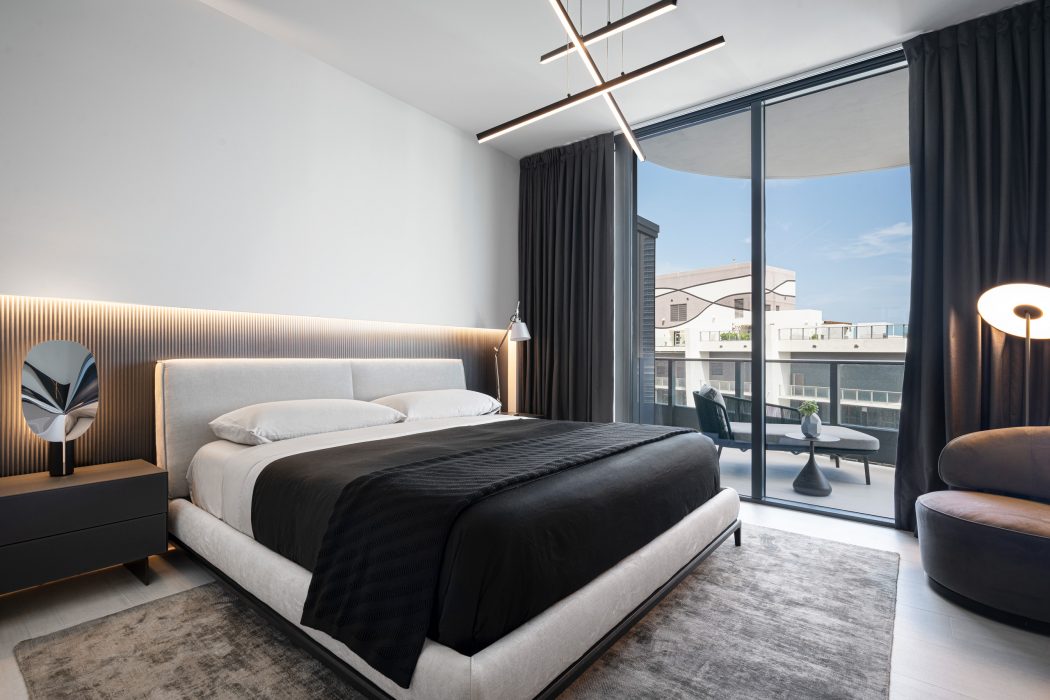 Professional High-end Interior Design composition of all black-and-white bedroom, white bed with white sheets and black duvet cover, dark gray rug, dark gray round lounge chair, dark gray heavy curtains, headboard wall with half bottom part with mettalic black stripped wallpaper illuminated with indirect LED lights, and glossy chrome leaf shape table lamp